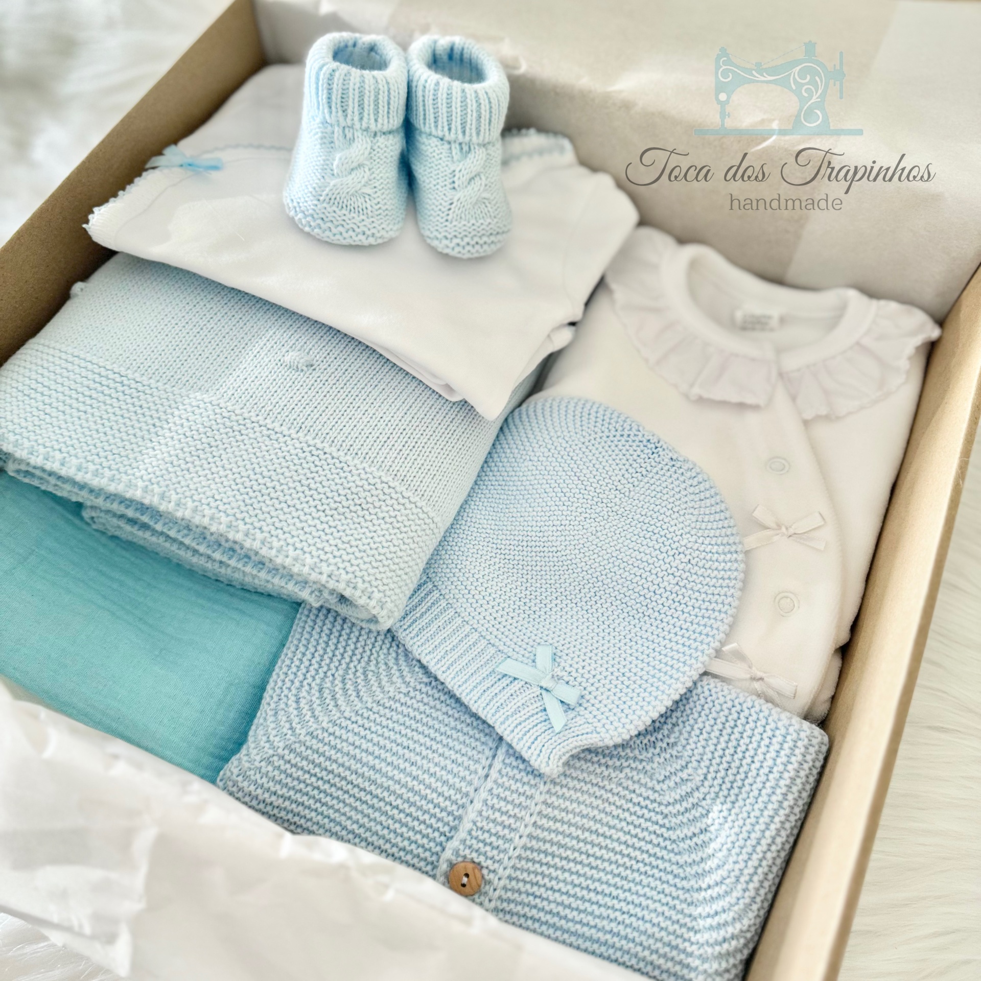 Conjunto de roupa de bebé azul claro e branco numa caixa aberta com botinhas, gorro, manta e body