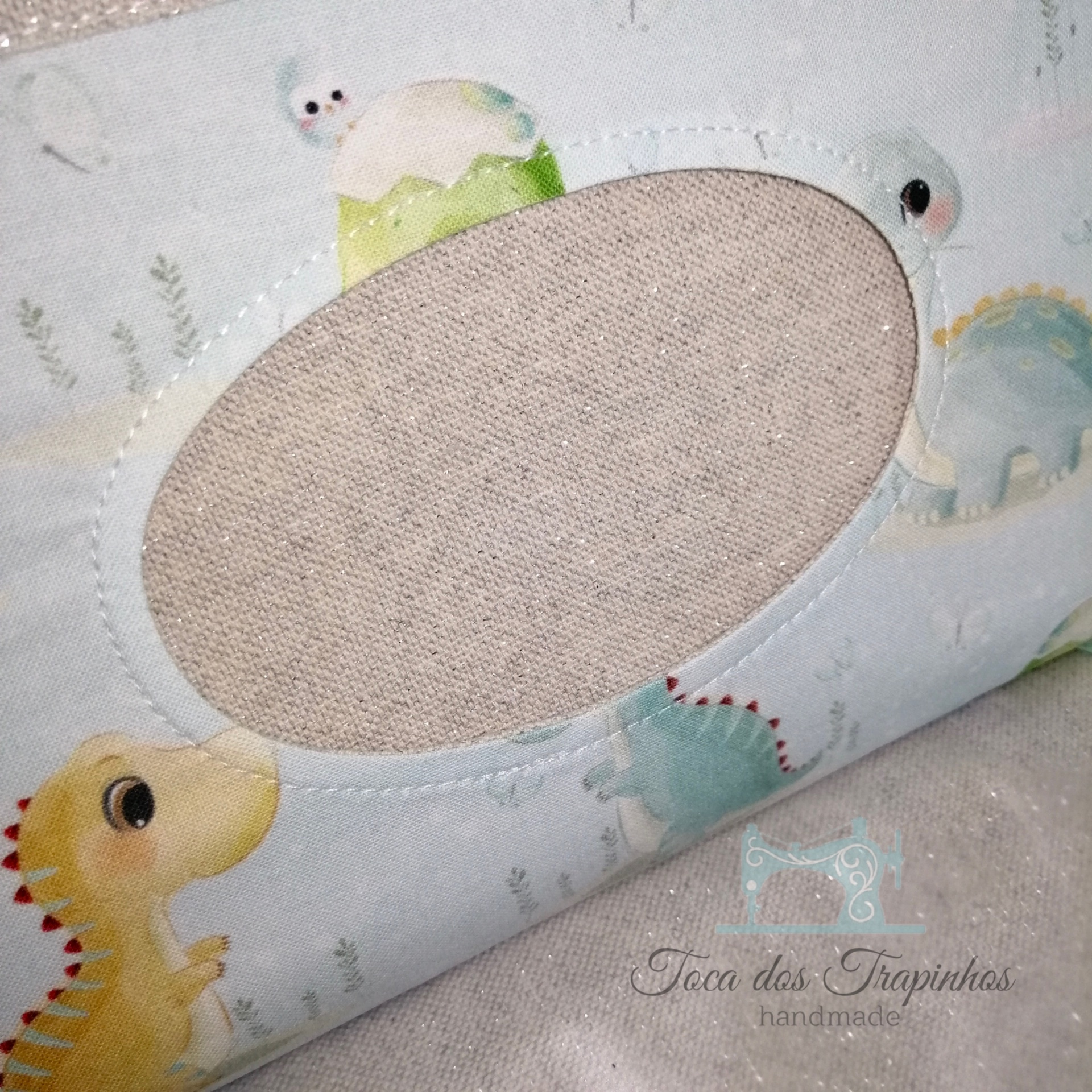 Almofada azul clara com dinossauros coloridos e abertura oval central em tecido bege