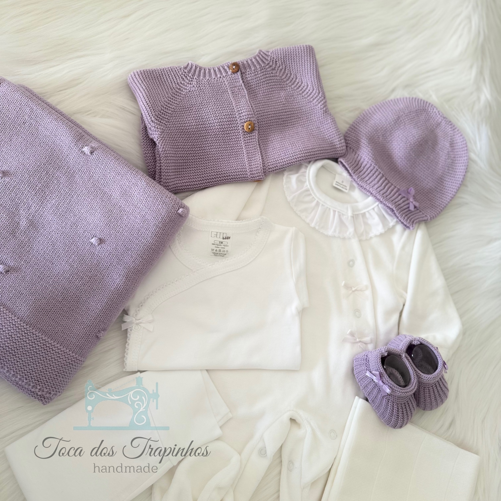 Conjunto de roupa de bebé branco e lilás com manta, camisola, gorro e sapatinhos sobre fundo de pelo branco