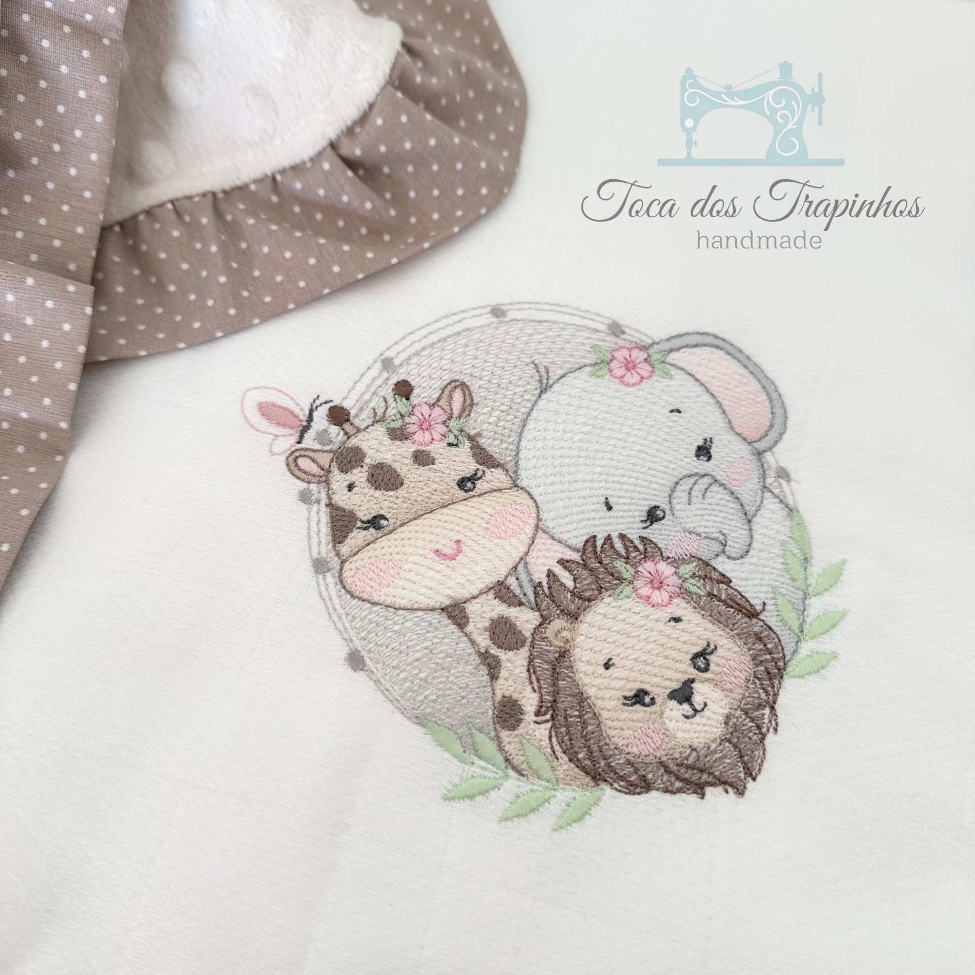 Manta branca com bordado de girafa, elefante e leão, tecido castanho com bolinhas brancas na bainha