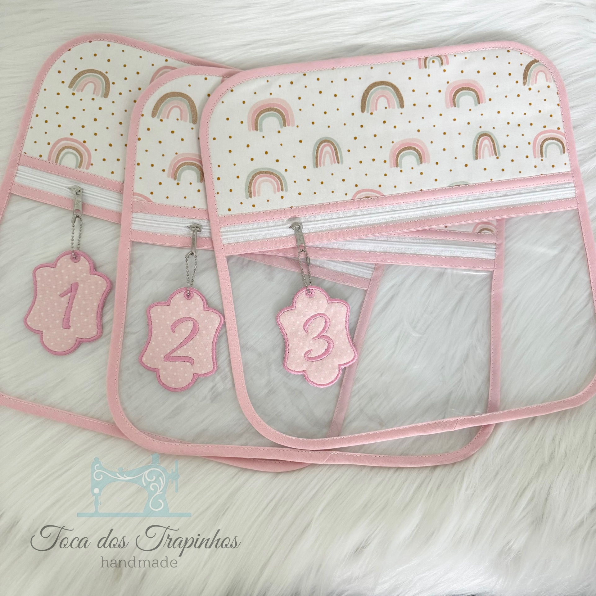 Três organizadores transparentes com bordas e estampas rosa e chaveiros numerados em fundo branco peludo