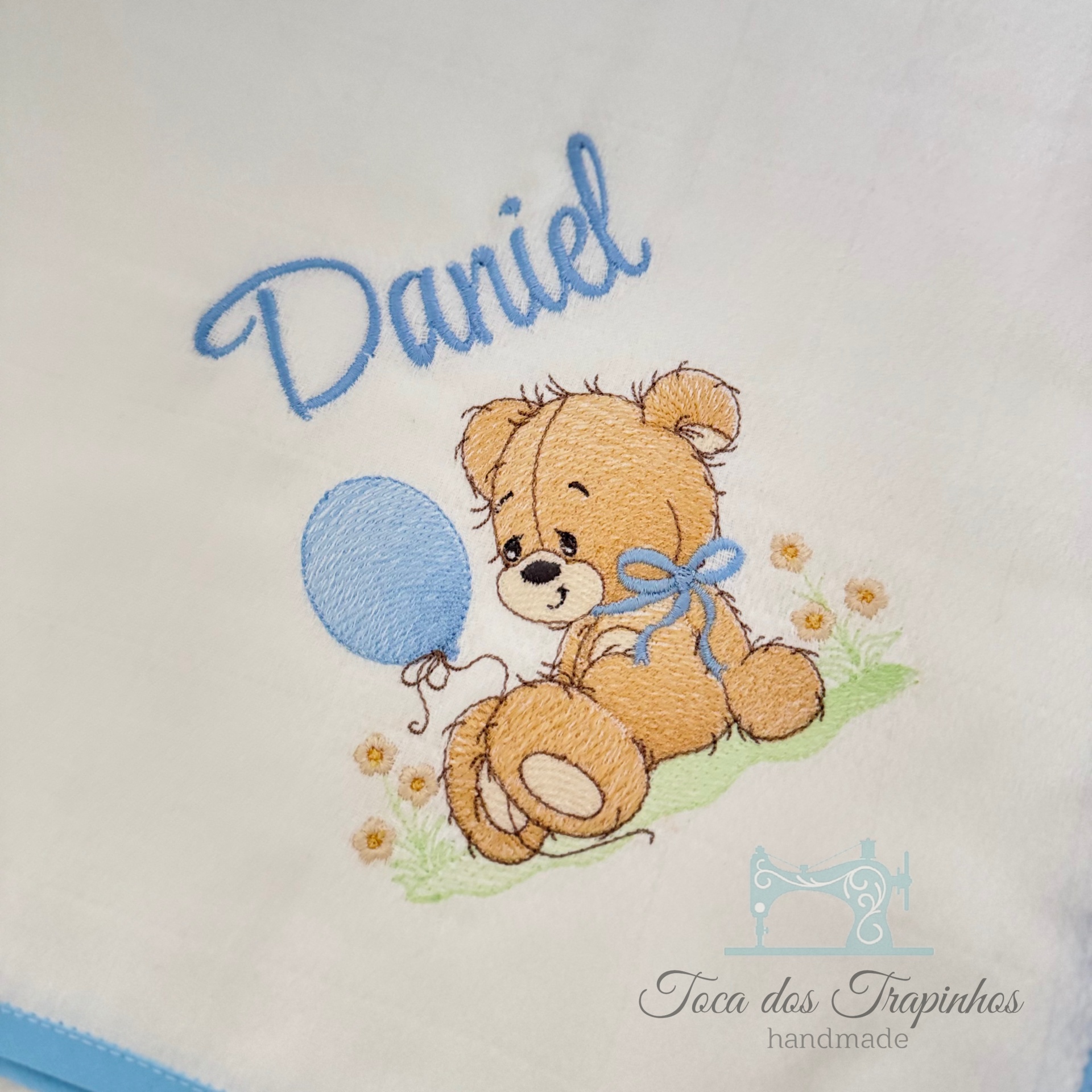 Toalha branca bordada com ursinho castanho e balão azul, texto Daniel e logo Toca dos Trapinchos