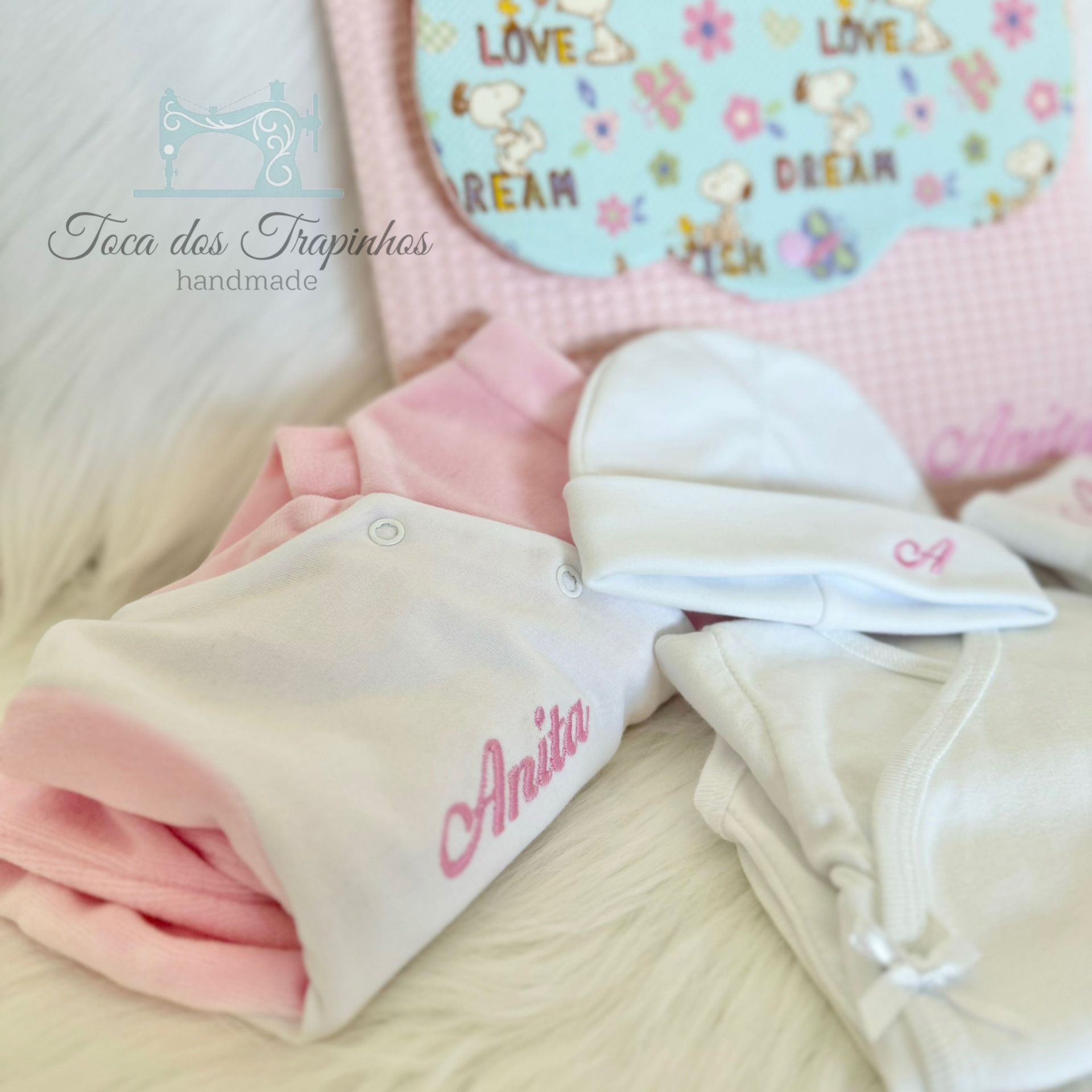 Roupas de bebé rosa e branco com nomes bordados, pano azul decorativo com Snoopy