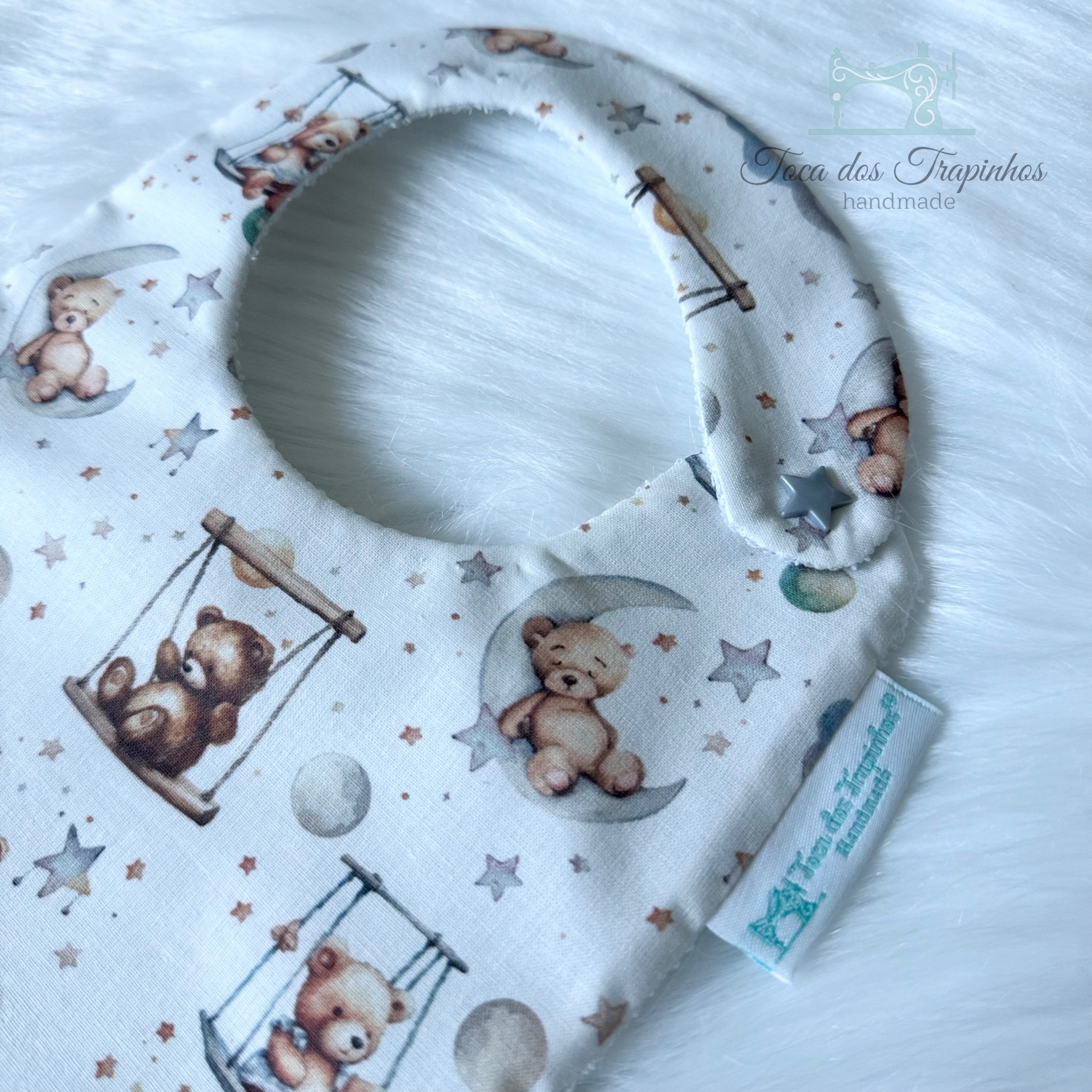 Babete para bebé com padrão de ursos em balanço, estrelas e bolas, etiqueta azul, fundo branco.