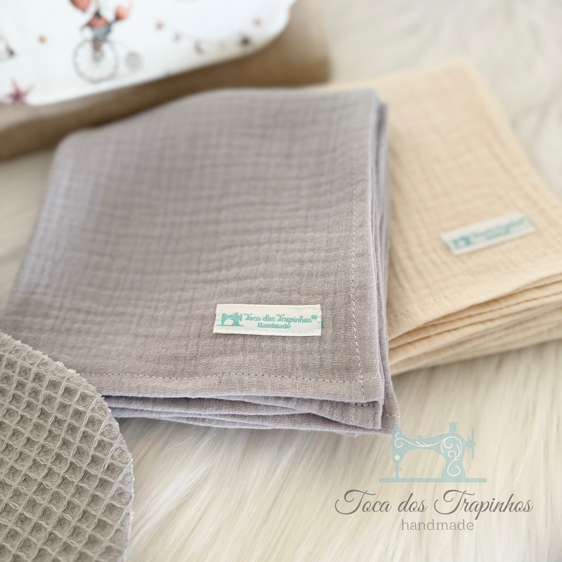Mantinhas de tecido cinza e bege com etiqueta 'Toca dos Trapinhos Handmade' sobre superfície branca felpuda