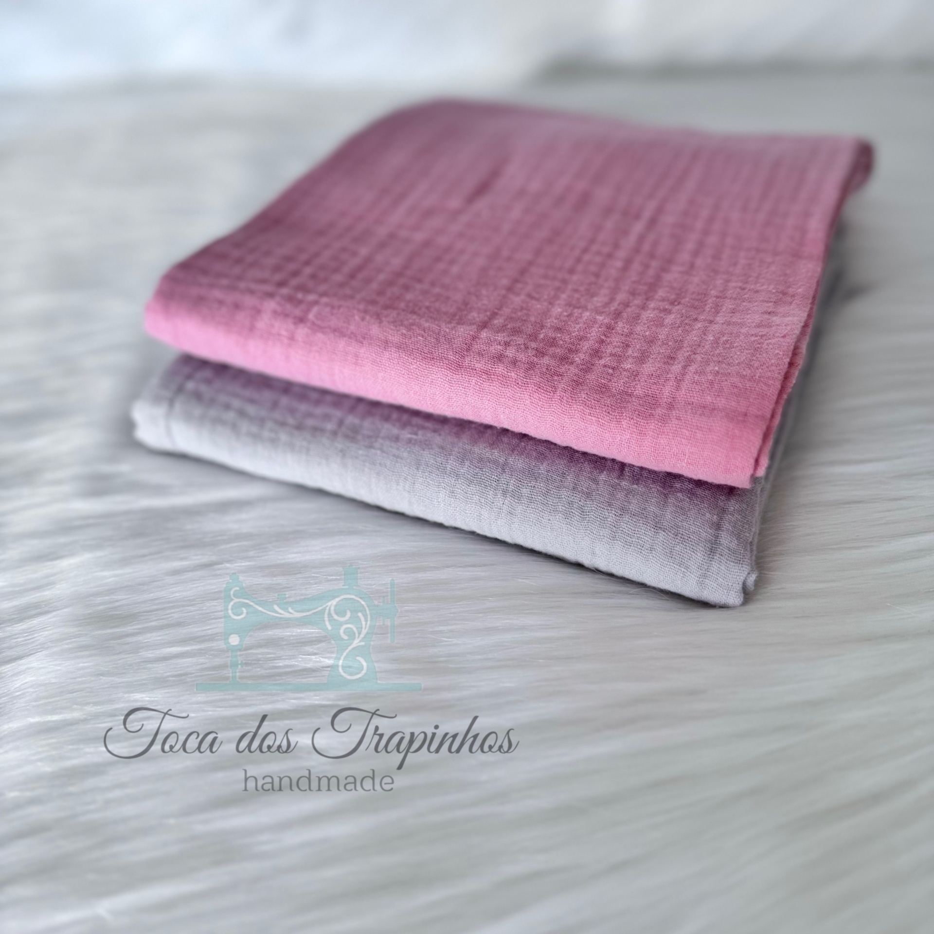 Dois tecidos dobrados rosa e cinzento claro sobre superfície branca com texto 'Toca dos Trapinhos handmade'.