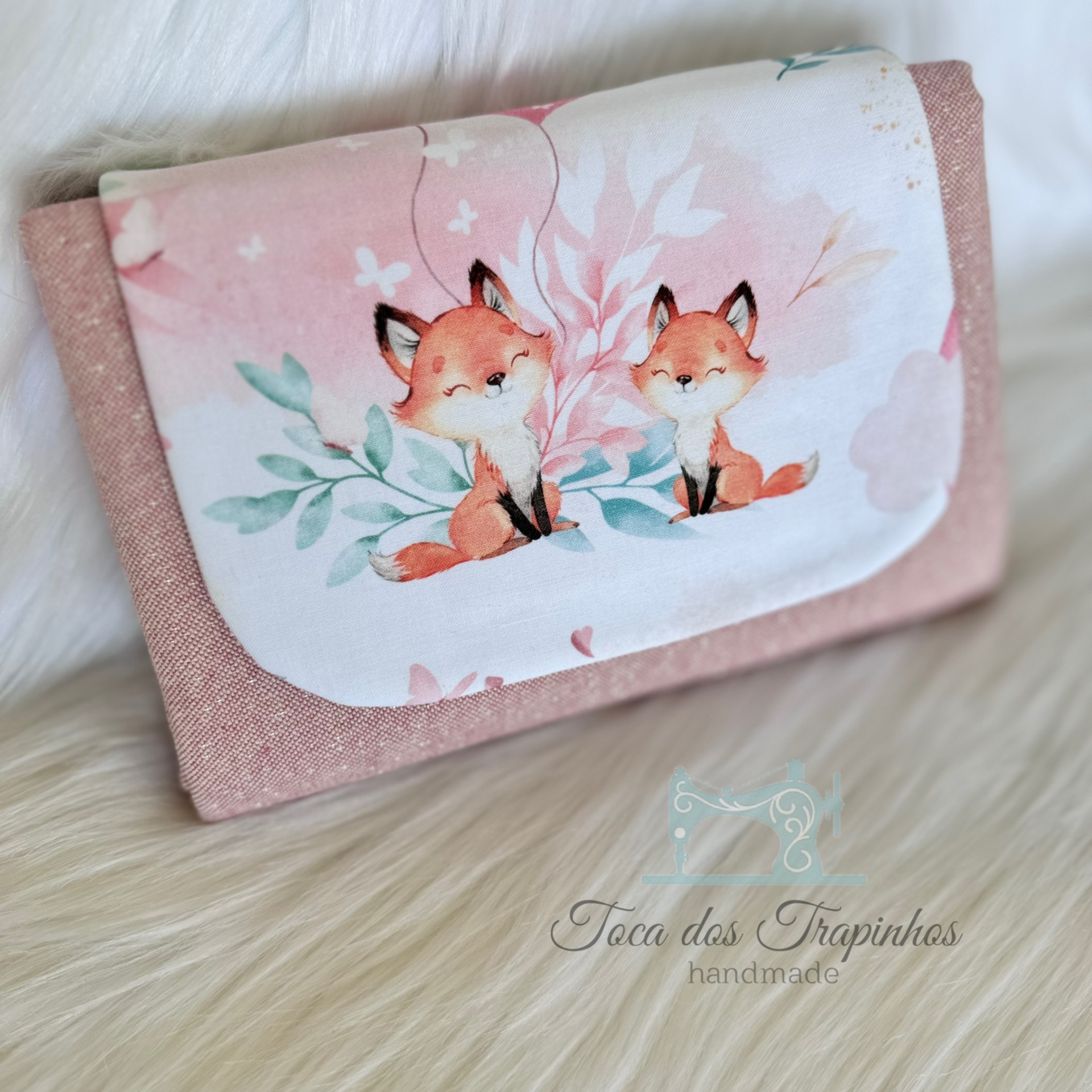 Carteira rosa com estampa de raposas e folhas sobre superfície branca peluda