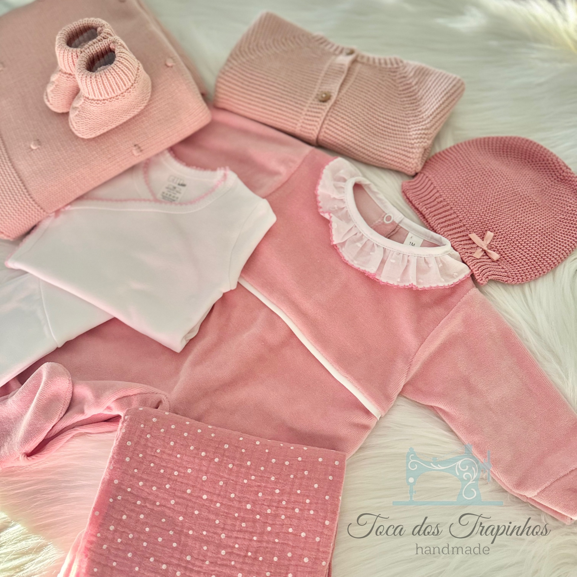 Conjunto de roupa de bebé rosa com vários itens em malha e tecidos suaves, incluindo sapatinhos, casaco, body, vestido, touca e manta, em fundo branco