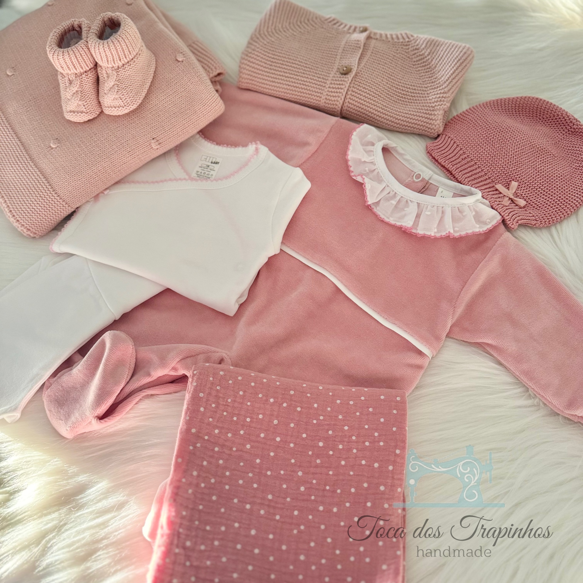 Conjunto de roupa de bebé rosa e branco com várias peças e manta, sobre superfície branca fofa