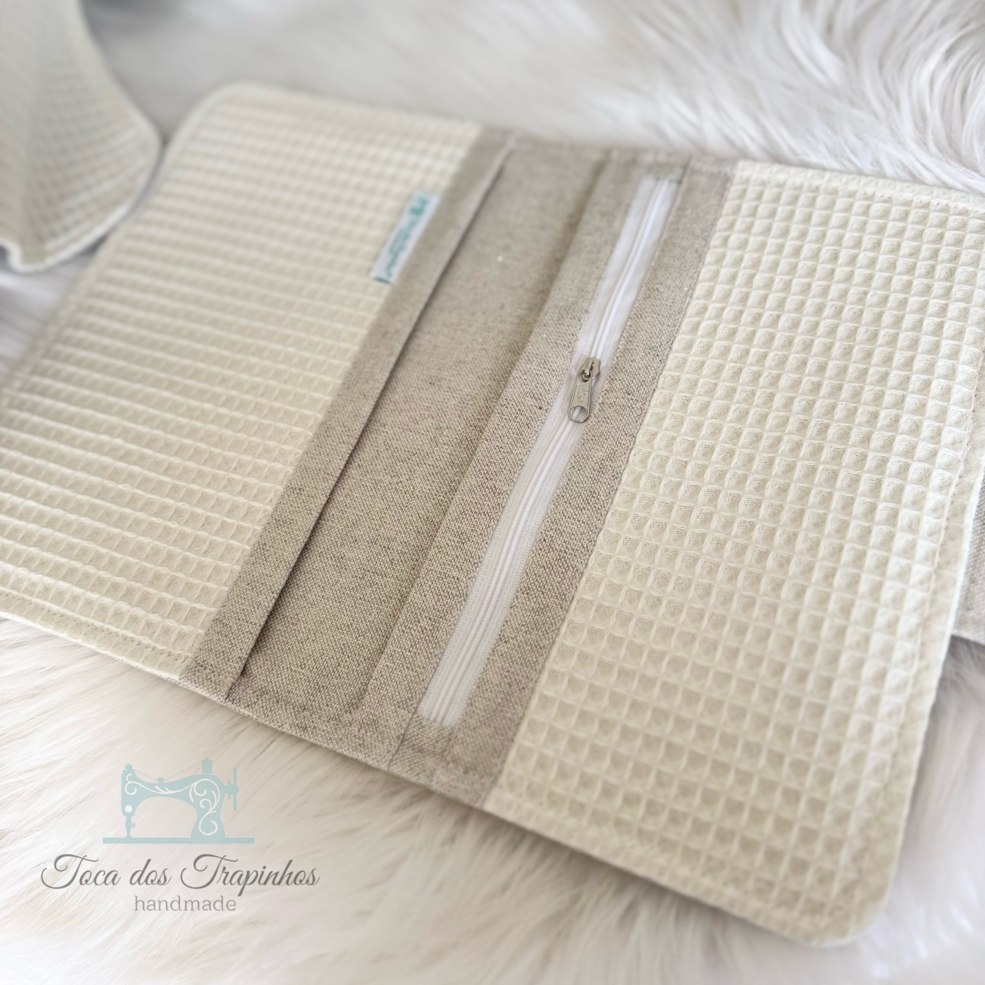 Carteira em tecido bege com zíper e padrão waffle, sobre superfície branca felpuda.