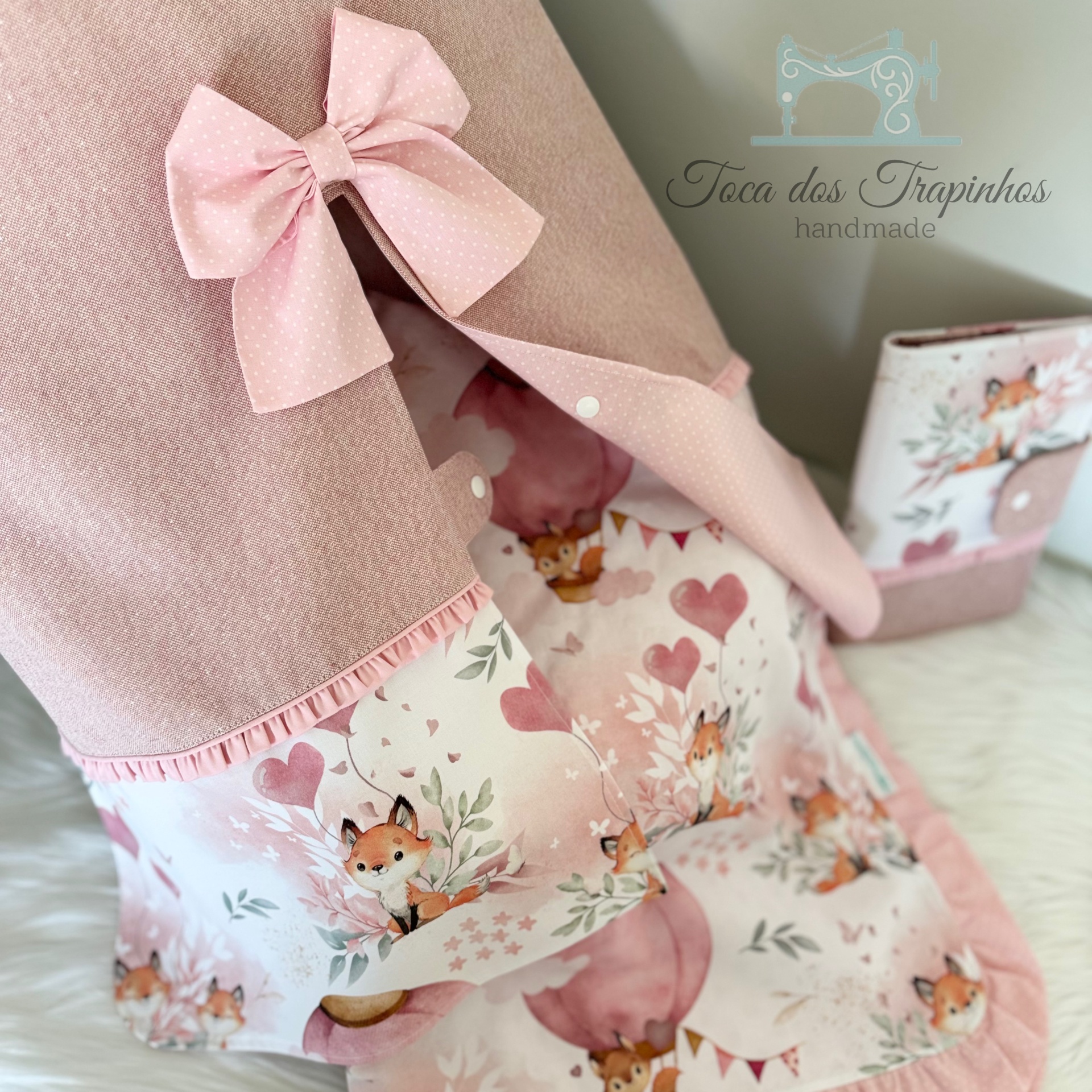 Produto têxtil rosa com padrão de raposas e laço, próximo a caixa rosa com padrão floral.