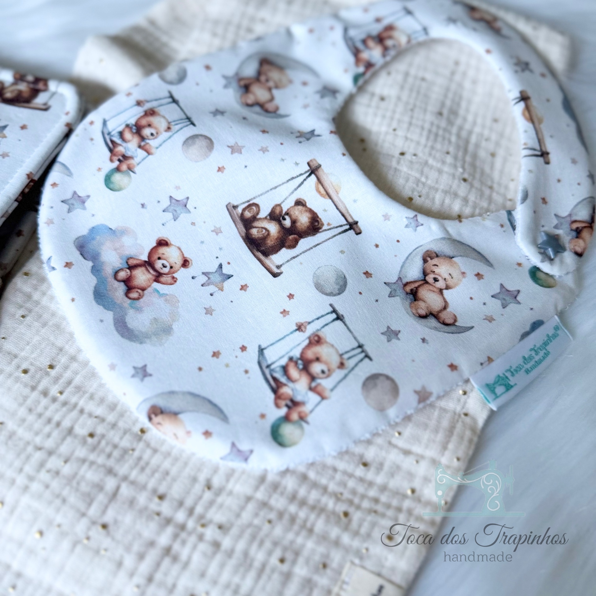 Babete infantil com padrão de ursinhos castanhos, estrelas e luas em tecido branco.