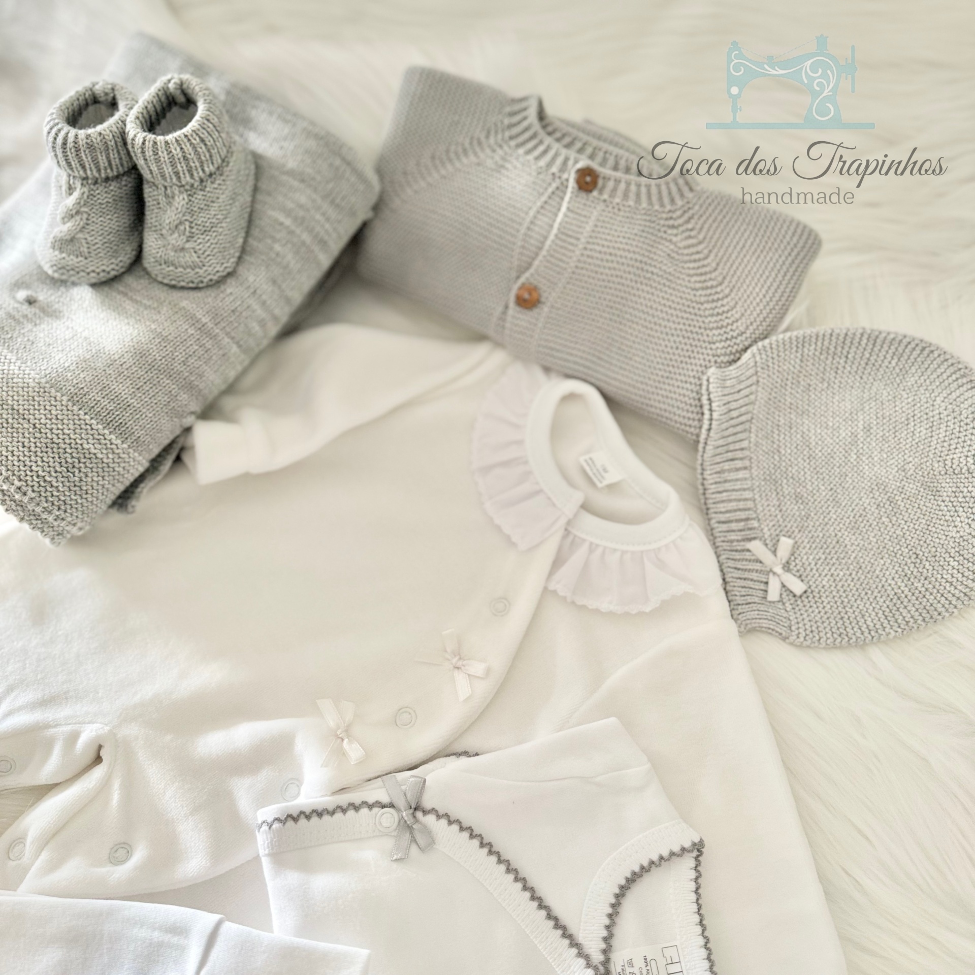 Conjunto de roupa de bebé branca e cinza com bodie, casaco, gorro, sapatinhos e manta sobre superfície branca