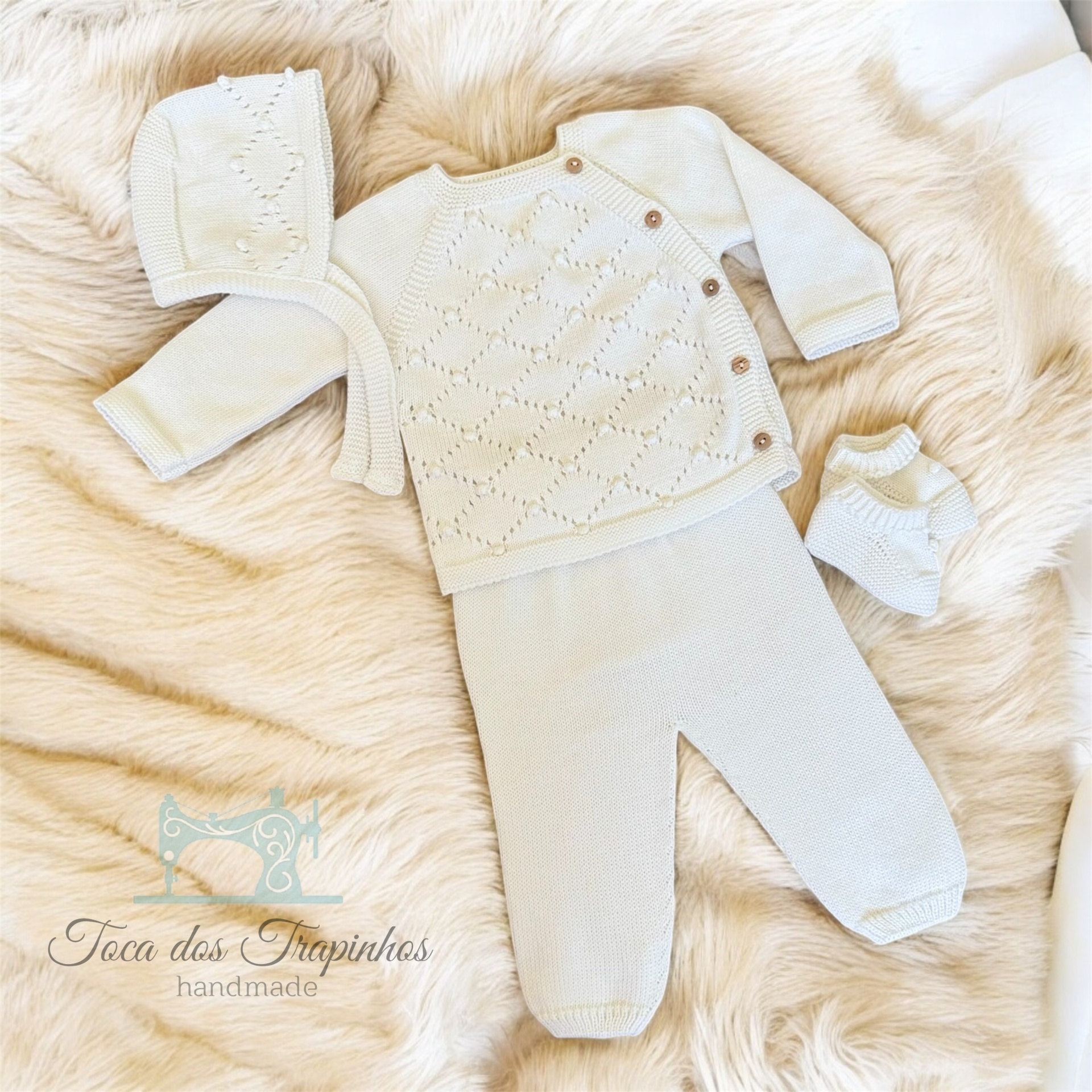 Conjunto de bebé branco de malha com camisola, calças, gorro e botins sobre fundo de pelo bege.