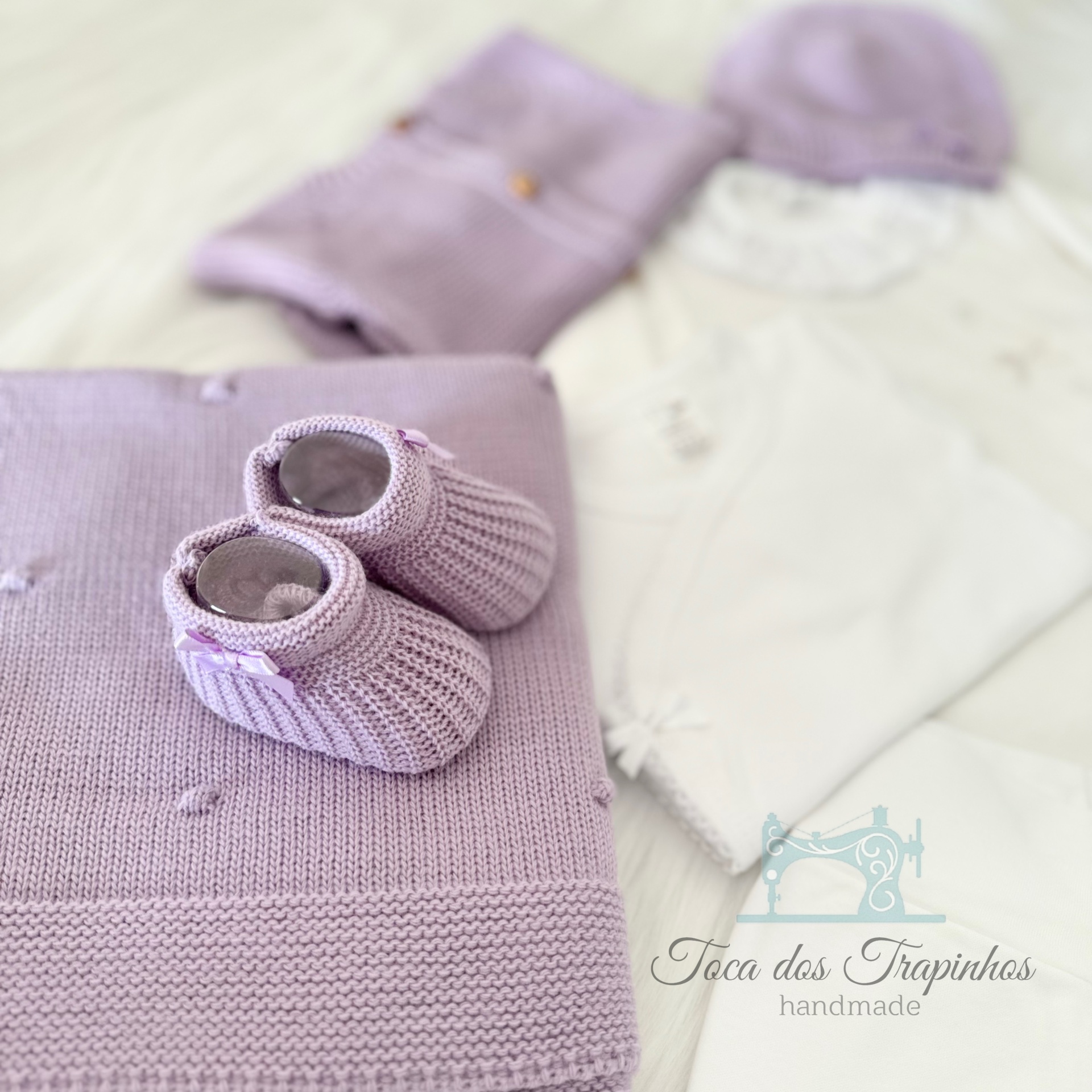 Conjunto de roupas e acessórios de bebé em lilás e branco, com sapatinhos de malha sobre manta lilás.