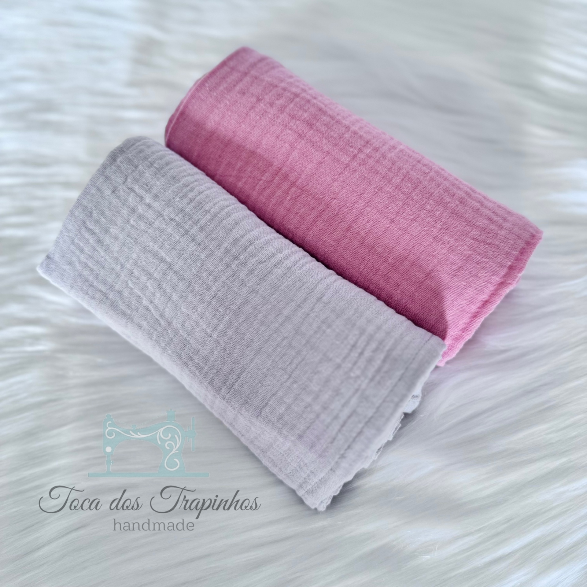 Dois panos de algodão rosa e cinza com textura canelada sobre superfície branca macia com logo 'Toca dos Trapinhos handmade'.