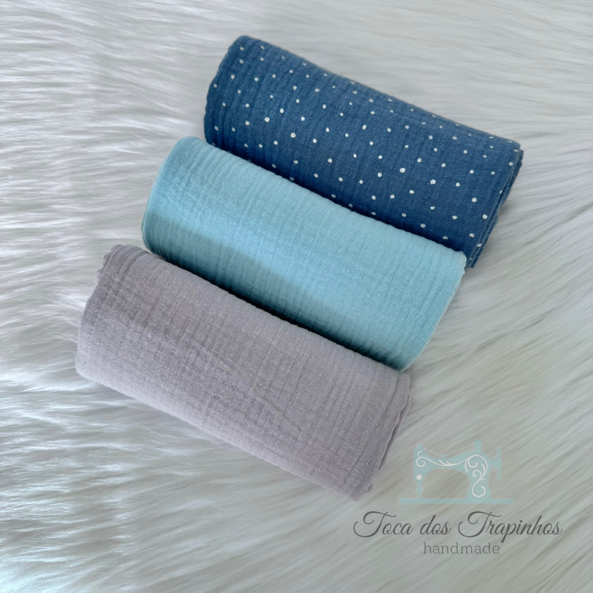 Três panos de tecido enrolados em azul escuro com bolinhas brancas, azul claro e cinzento sobre superfície branca felpuda com logo
