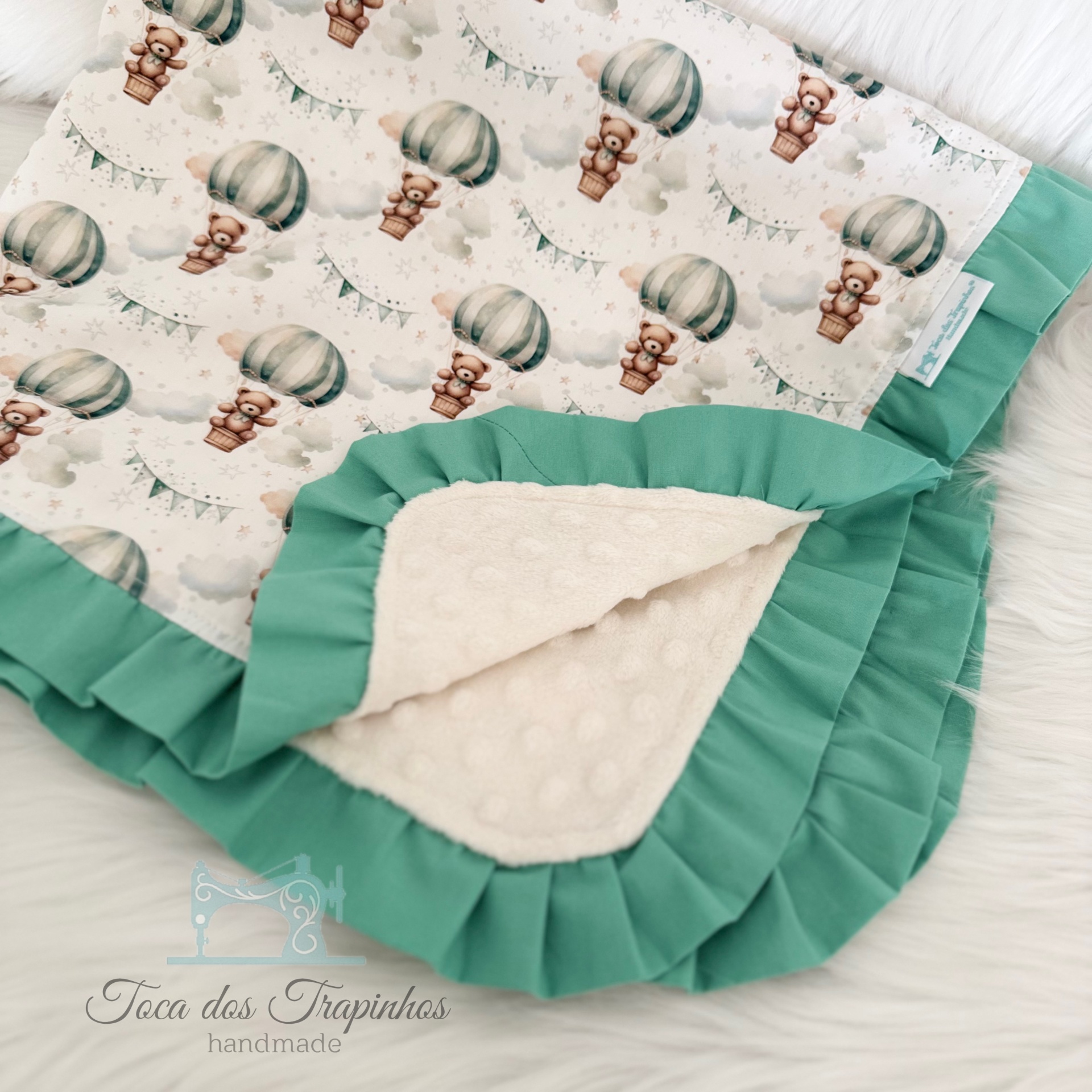 Manta para bebé com padrão de balões de ar quente e ursinhos, folhos verdes e tecido interior branco texturado.