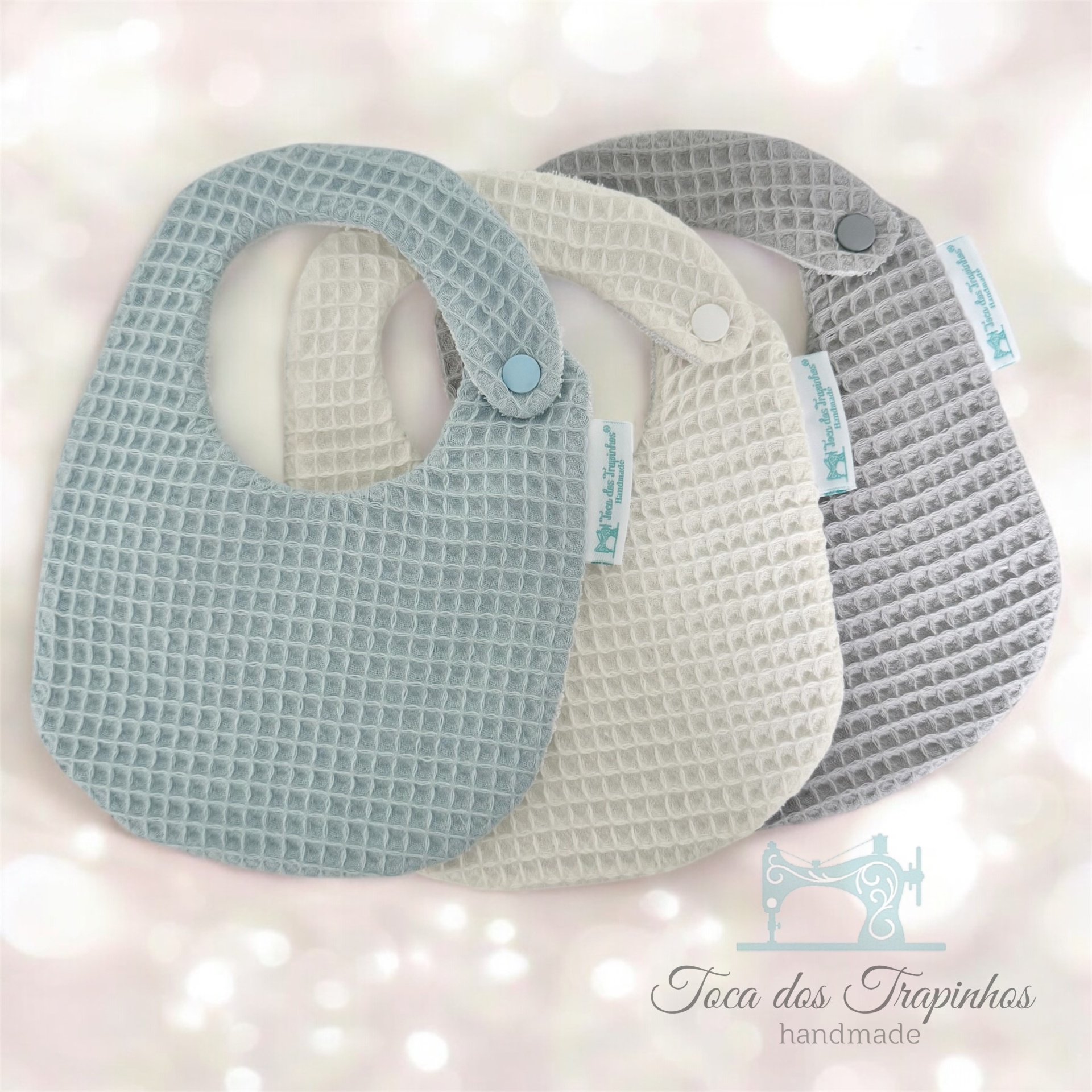 Três babetes em tecido com padrão de favo, azul claro, creme e cinza, com fecho de pressão