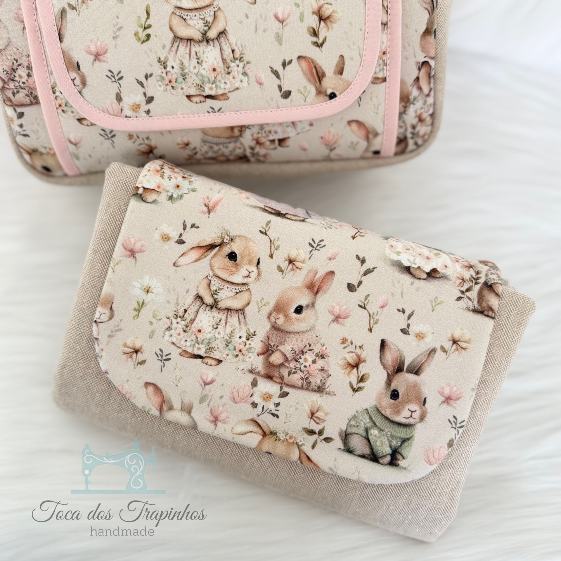 Carteira em tecido bege claro com padrão de coelhos vestidos e flores, junto com bolsa rosa de padrão semelhante