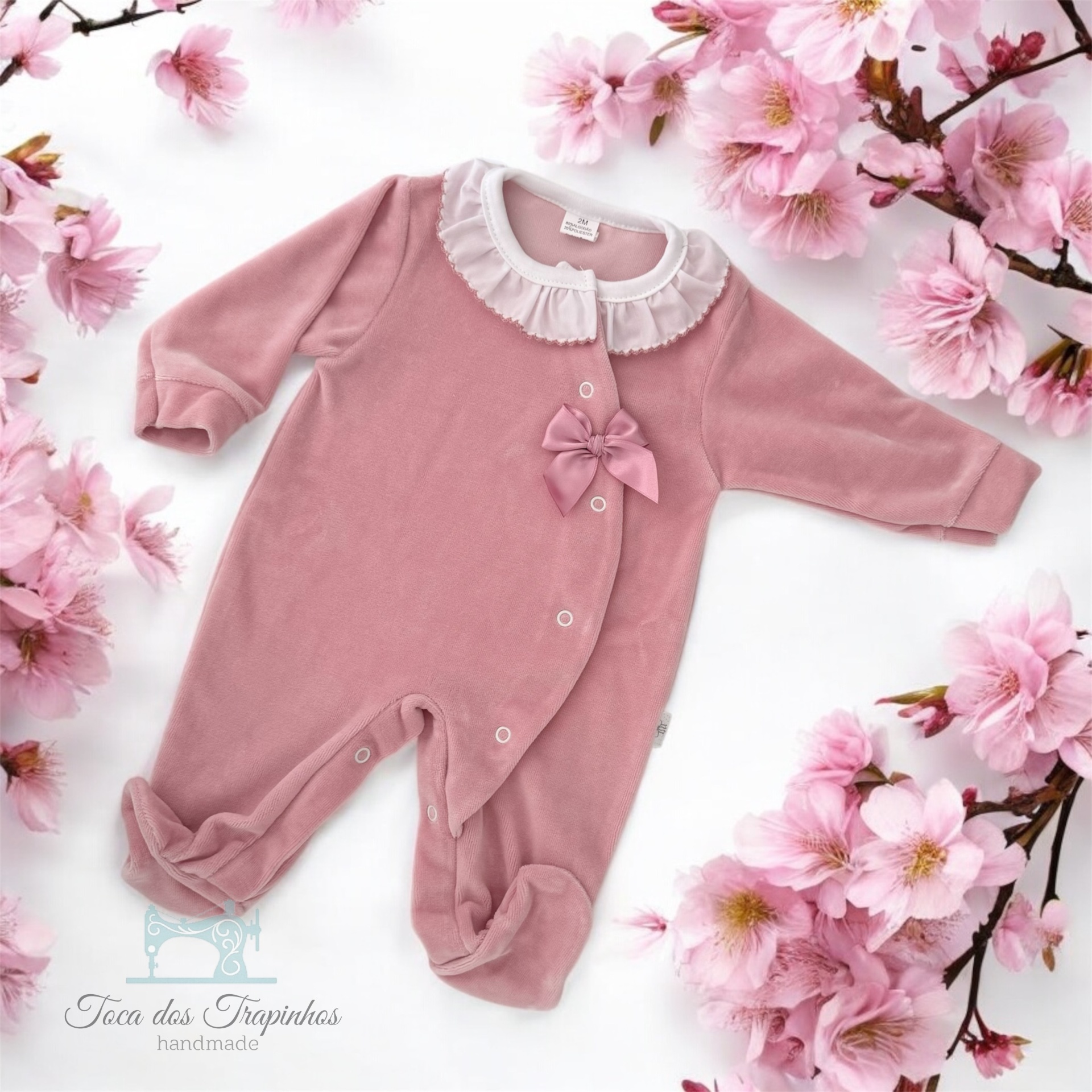 Macacão de bebé cor-de-rosa com gola branca e laço, rodeado por flores cor-de-rosa