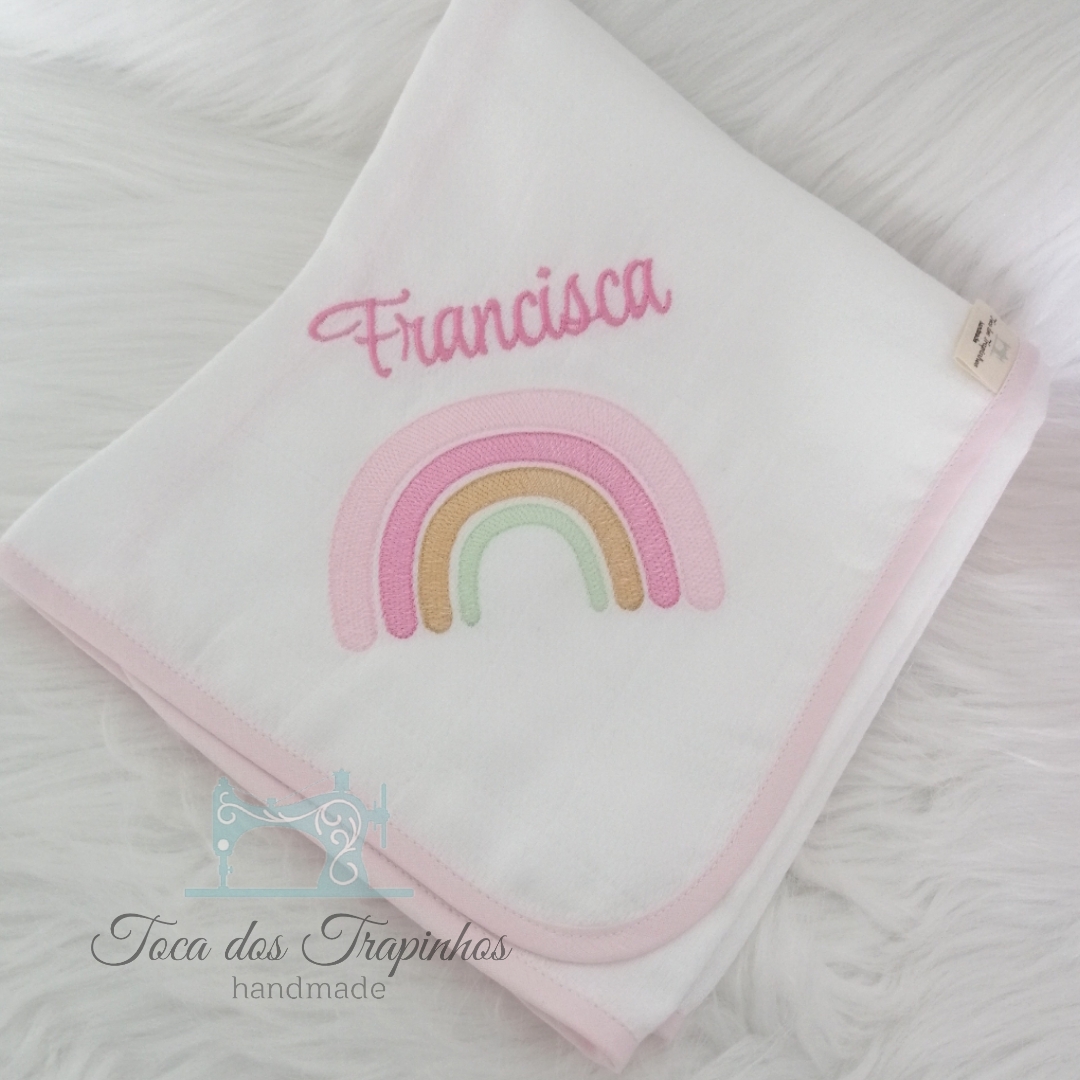 Manta branca com bordas cor de rosa, bordado de arco-íris colorido e o nome Francisca