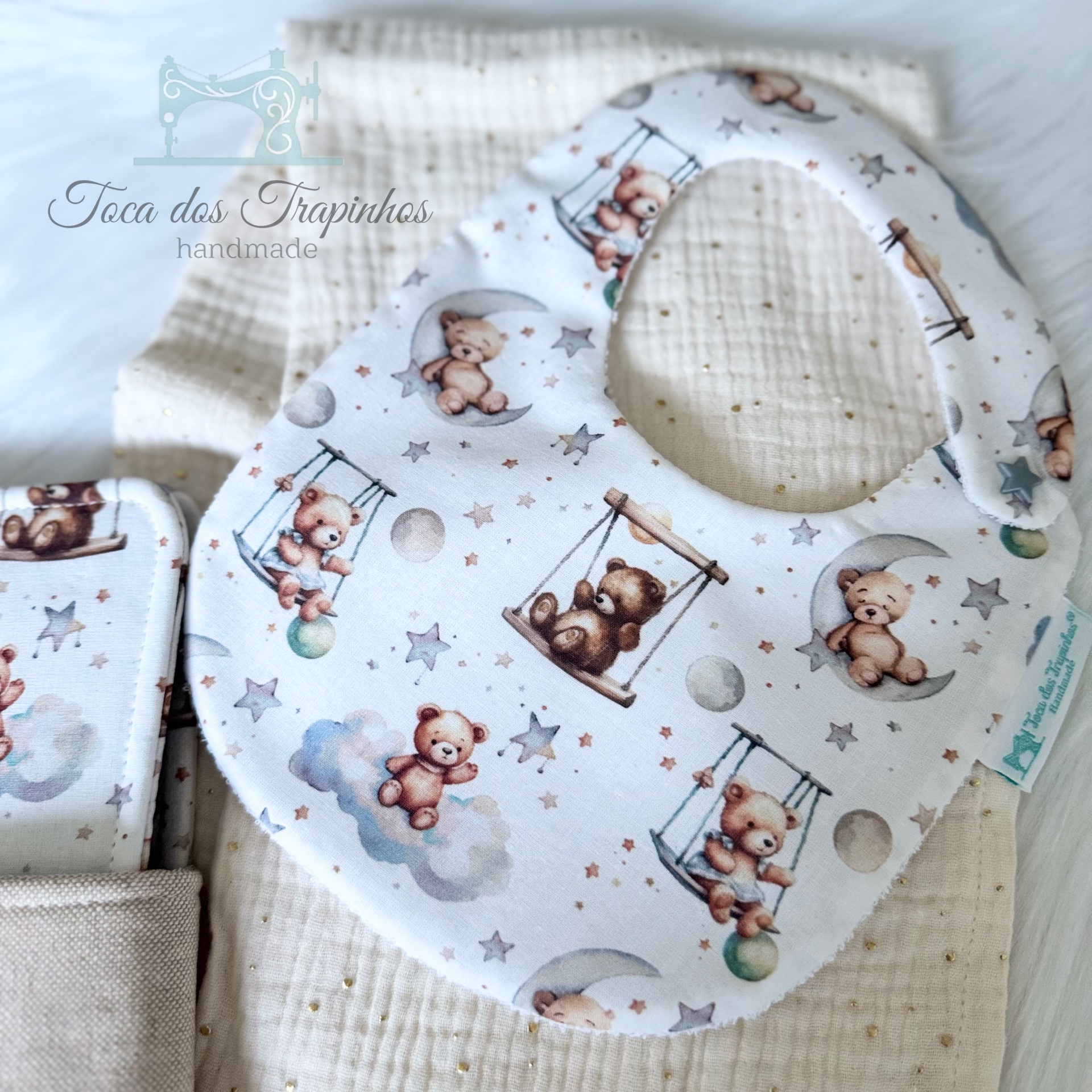 Babete branco com padrão de ursinhos em balanços, estrelas e luas sobre tecido bege texturado