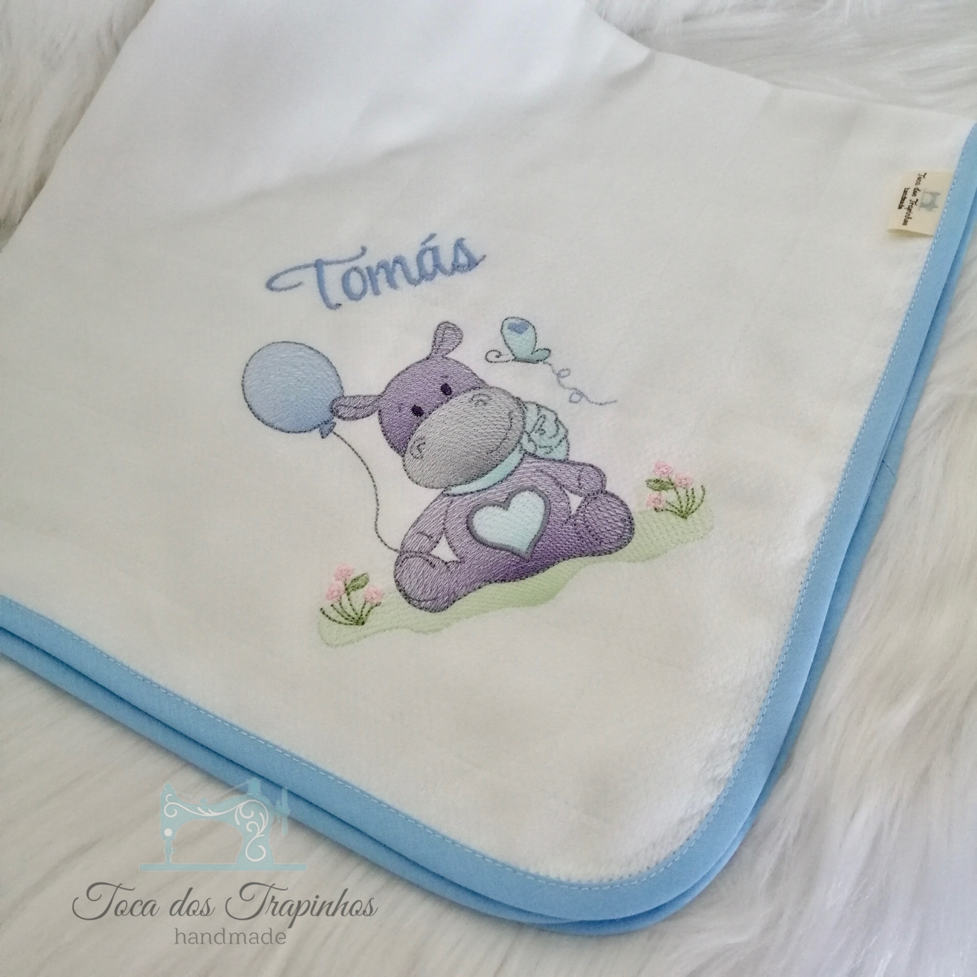 Manta branca para bebé com borda azul e bordado de hipopótamo lilás com balão e passarinho, nome Tomás em azul
