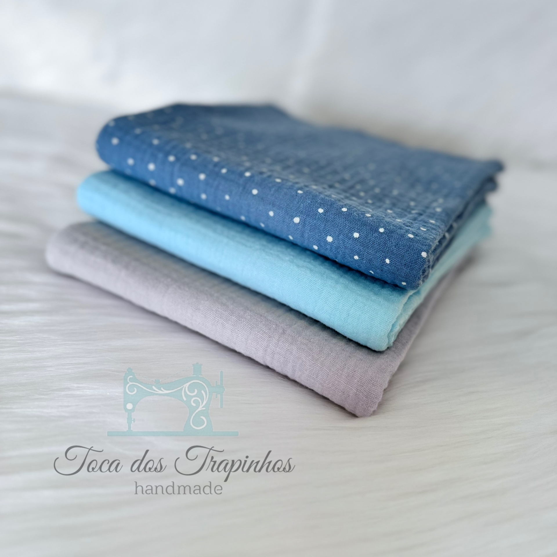 Três tecidos azuis dobrados com padrão de bolinhas e logotipo Toca dos Trapinhos
