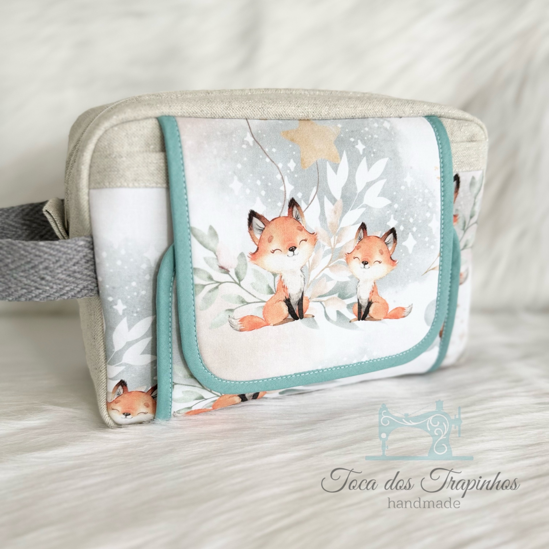 Necessaire bege com estampa de duas raposas sorridentes em tecido com folhas e estrelas, bordas azuis e alça cinza.