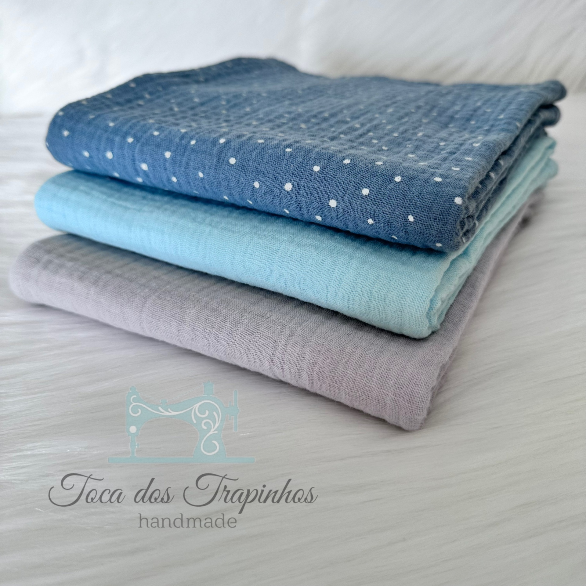 Três tecidos dobrados empilhados em azul com bolinhas, azul claro e lilás sobre fundo branco.