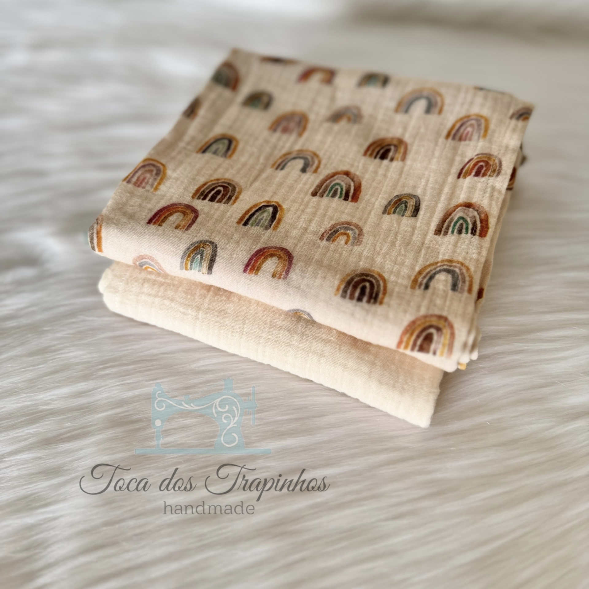 Duas mantas de tecido bege com padrões de arcos-íris e lisa sobre fundo branco felpudo.