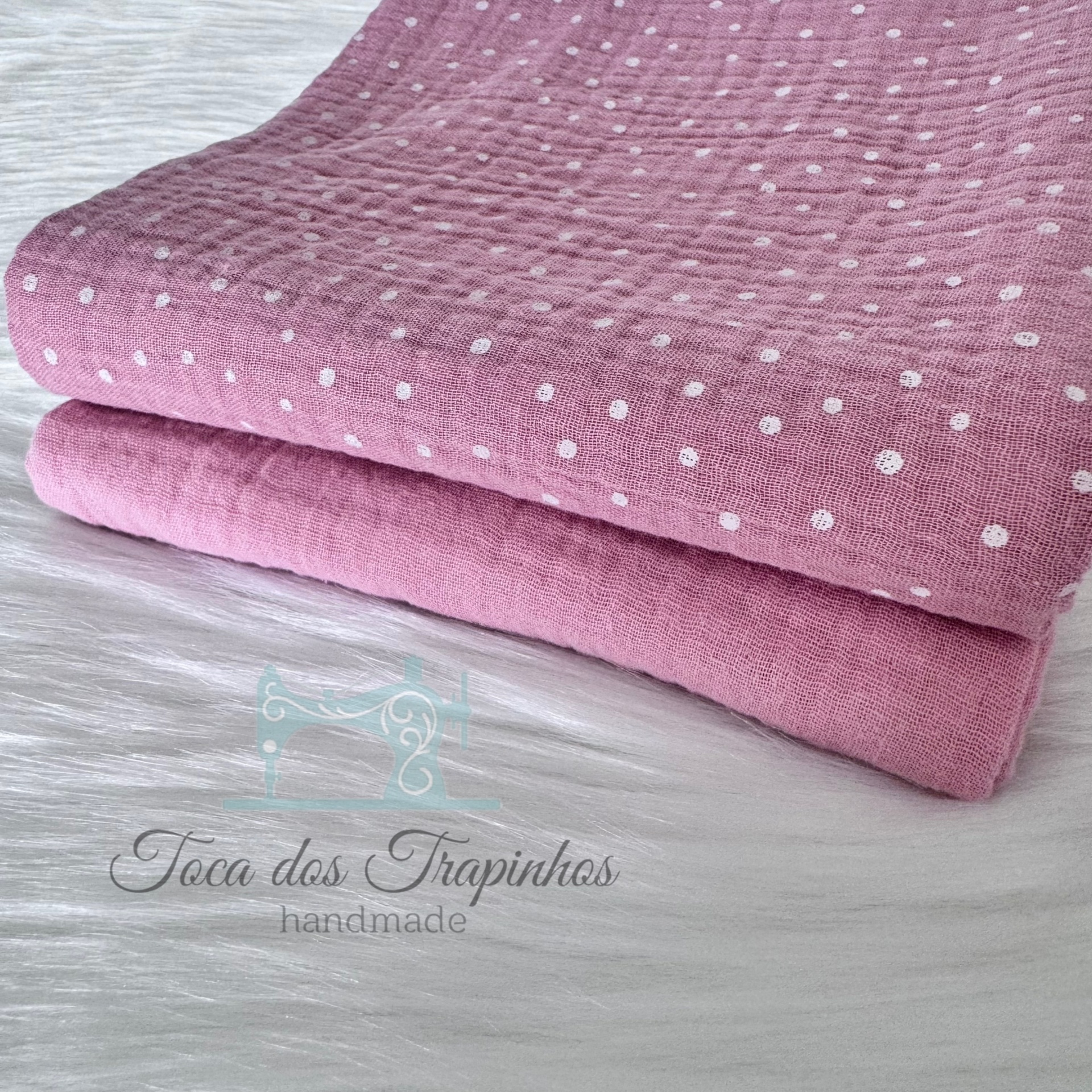 Dois tecidos rosa, um com padrão de pontos brancos e outro liso, dobrados sobre superfície branca com logo da marca.
