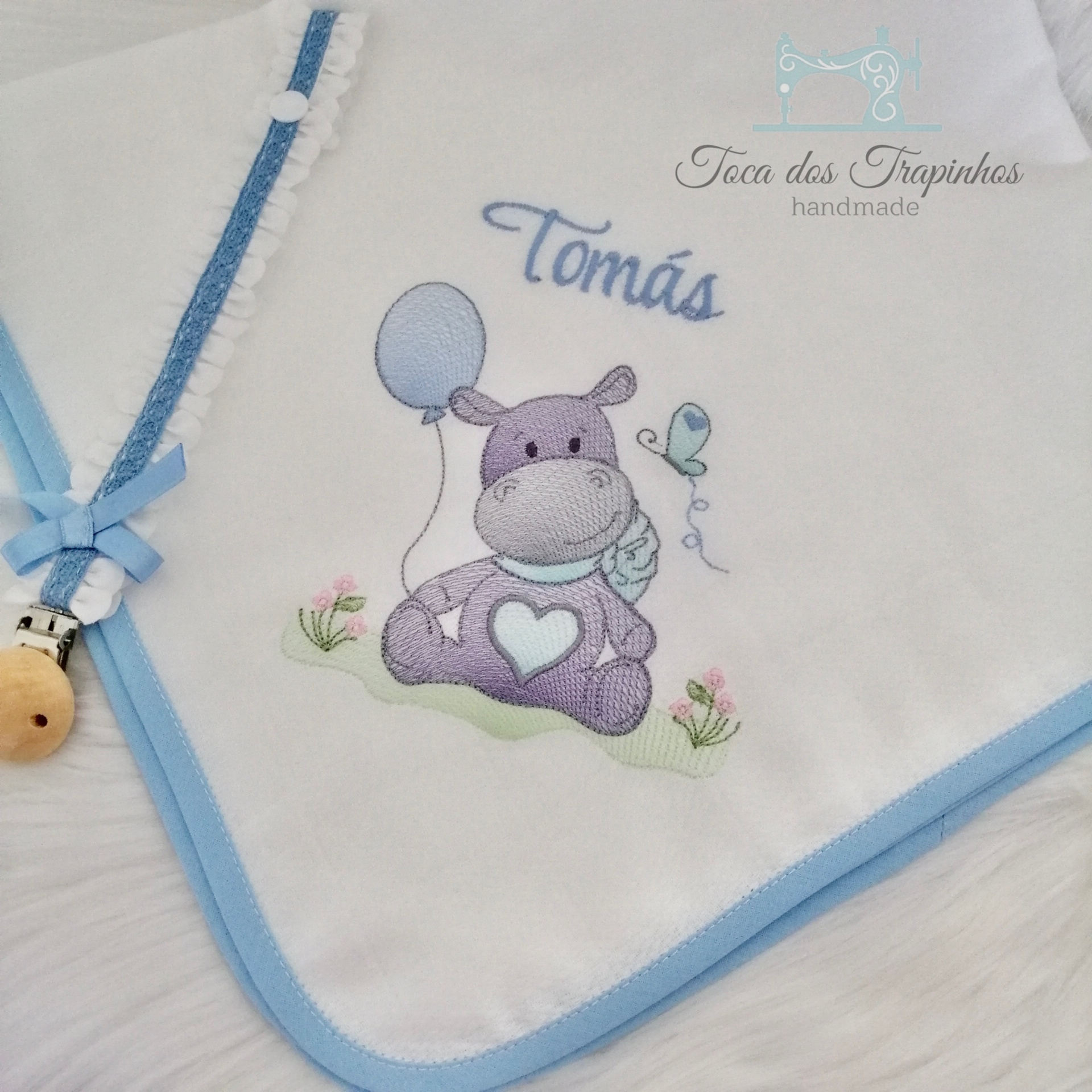 Fralda branca com bordas azuis, hipopótamo lilás, nome 'Tomás' bordado, fita azul com presilha de madeira