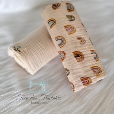 Tecidos infantis em gaze, um creme liso e outro creme com arcos-íris, sobre superfície branca felpuda.