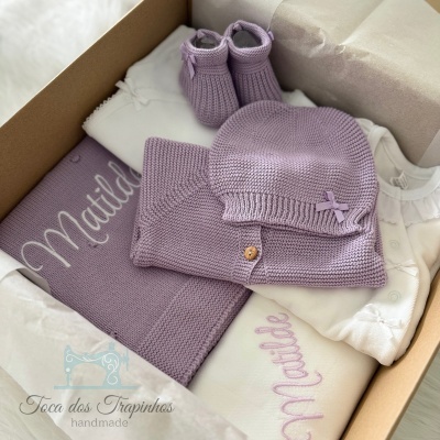 Conjunto de roupa de bebé lilás e branco numa caixa com manta bordada e sapatinhos de malha.
