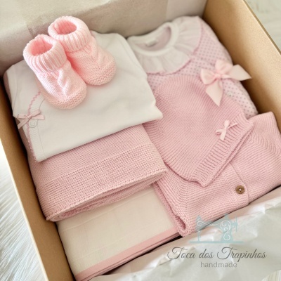 Conjunto de roupa de bebé cor-de-rosa e branco com sapatinhos numa caixa de cartão.