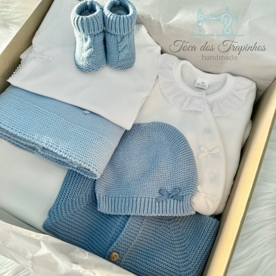 Conjunto de roupa de bebé azul e branco com gorro, botins, casaco e body numa caixa com papel branco