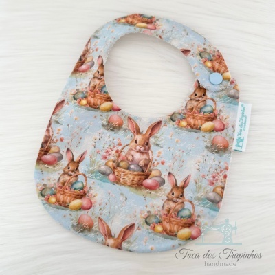 Babete para bebé com padrão de coelhos, ovos coloridos e fundo azul claro