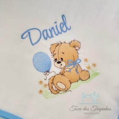 Toalha branca bordada com ursinho castanho e balão azul, texto Daniel e logo Toca dos Trapinchos