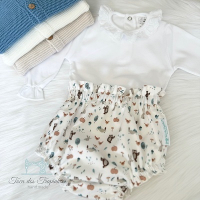 Conjunto de roupa de bebé com body branco de gola rendada e calções estampados, com peças de malha azul e bege ao lado, sobre superfície branca peluda.