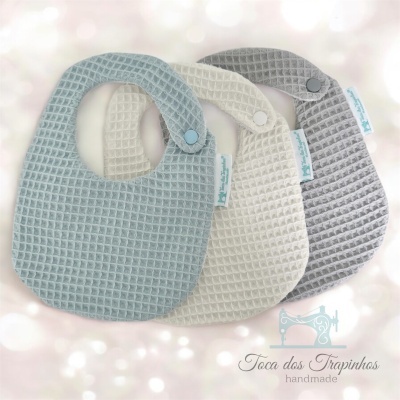 Três babetes em tecido com padrão de favo, azul claro, creme e cinza, com fecho de pressão