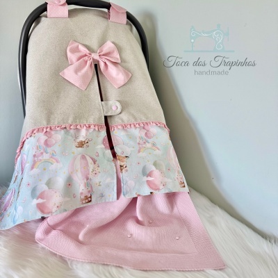 Capa para cadeirinha de bebé em tecido bege com laço cor-de-rosa e padrão de unicórnios e arco-íris