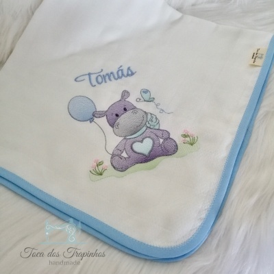 Manta branca para bebé com borda azul e bordado de hipopótamo lilás com balão e passarinho, nome Tomás em azul