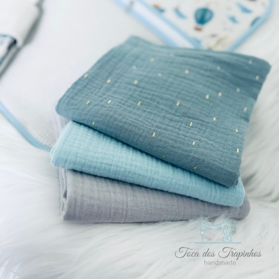 Três tecidos dobrados em tons de azul e cinza com pontos dourados sobre superfície branca macia