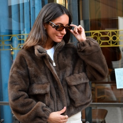 Mulher veste casaco de pelo castanho escuro com bolsos e óculos de sol.