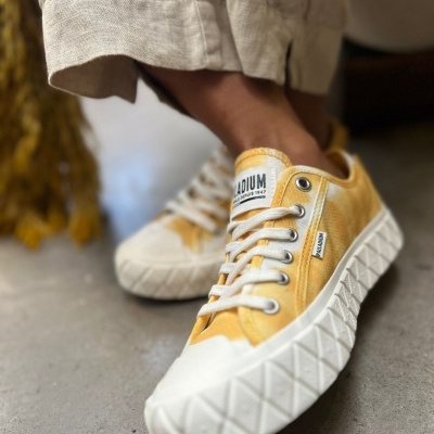 Ténis amarelos com sola branca texturizada e atacadores brancos em piso cinzento.