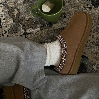Sapatos castanhos estilo mocassim com padrão em zig-zag usados com meias brancas e calças cinzentas junto a uma caneca verde com chá sobre um tapete.