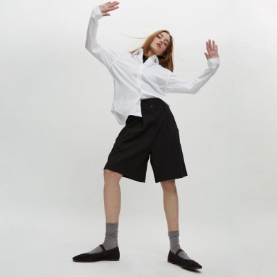Modelo com camisa branca, bermuda preta, meias cinza e sapatos pretos em fundo branco