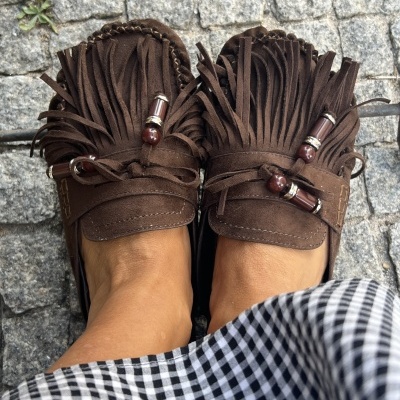 Mocassins castanhos em camurça com franjas e contas, pés nus, calçada de pedra